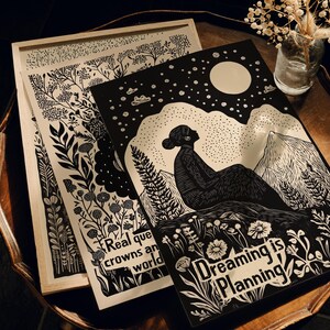 May include: Two black and white woodblock prints with a nature theme. The first print features a woman sitting on a mountain with a full moon and stars in the sky. The text reads "Dreaming is Planning". The second print features a variety of plants and flowers with the text "Real queens are crowned in a world".
