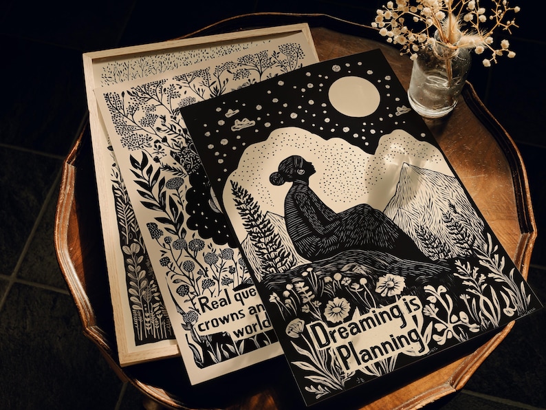 May include: Two black and white woodblock prints with a nature theme. The first print features a woman sitting on a mountain with a full moon and stars in the sky. The text reads "Dreaming is Planning". The second print features a variety of plants and flowers with the text "Real queens are crowned in a world".