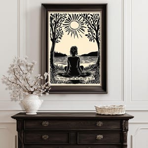 May include: Black and white framed art print featuring a person sitting by a lake, with trees on either side and a sun above. The text at the bottom reads, "The things we do do things to us."