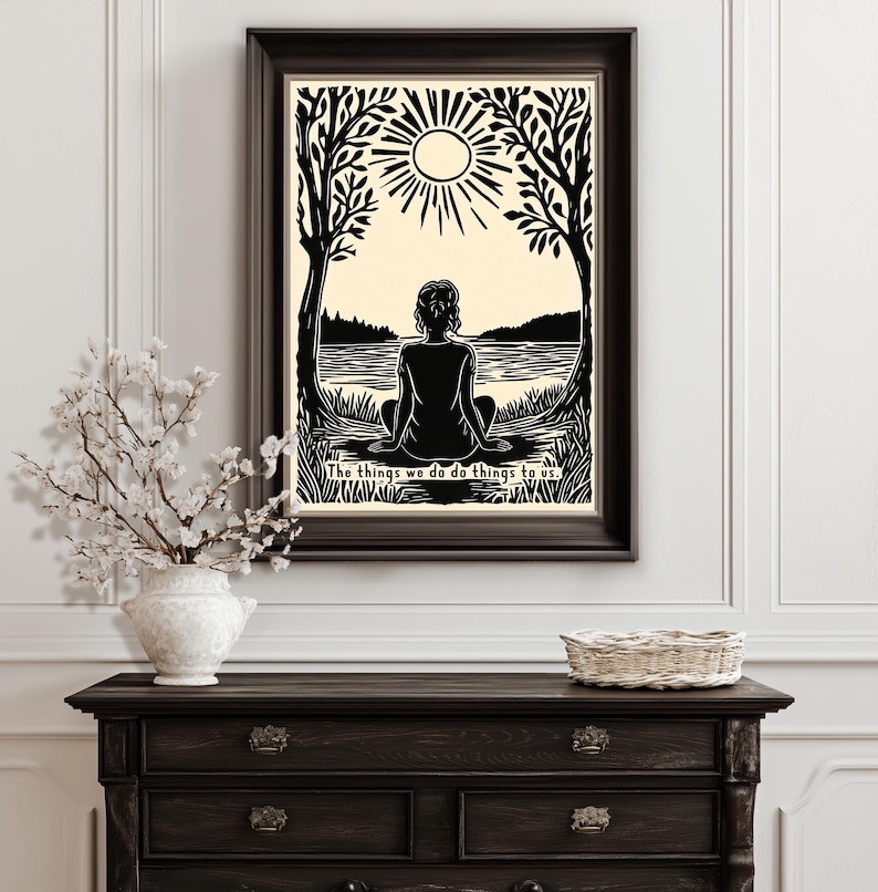May include: Black and white framed art print featuring a person sitting by a lake, with trees on either side and a sun above. The text at the bottom reads, "The things we do do things to us."