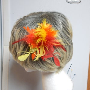 Może przedstawiać: Spinka do włosów z żółtych, pomarańczowych i czerwonych piór. Pióra są ułożone w kształt kwiatu.