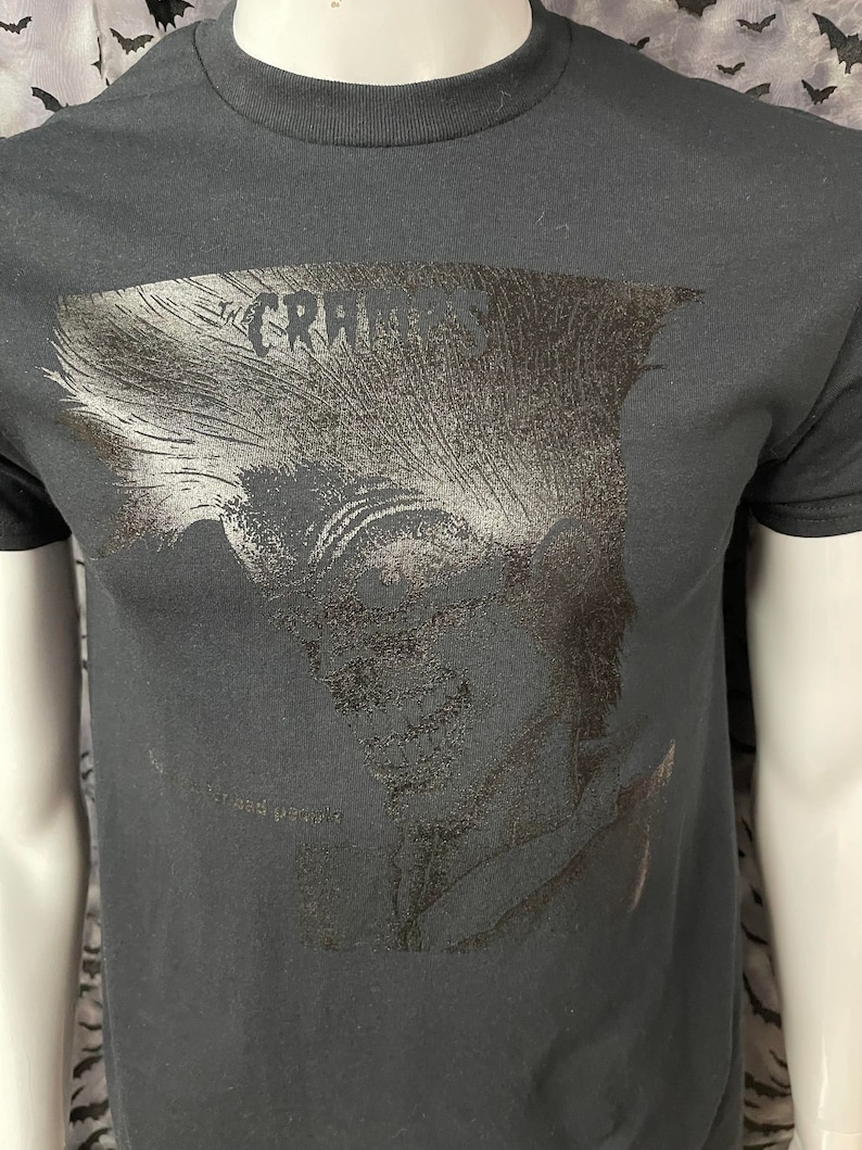 May include: Black t-shirt with a silver foil print of a person's face and the text "The Cramps" on the front.