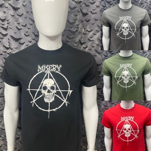 Op de afbeelding: Zwarte, groene en rode T-shirts met een wit schedel- en pentagram-ontwerp. De tekst "MISERY" is op elk shirt gedrukt.