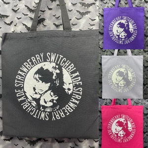 May include: Three tote bags with black handles and a white circle graphic featuring a woman's face and the text "Strawberry Switchblade" repeated around the circle. The bags are black, purple, and pink.