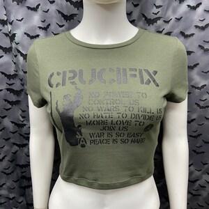 May include: A green cropped t-shirt with a black graphic print that says "CRUCIFIX" and includes the text "NO POWER TO CONTROL US, NO WARS TO KILL US, NO HATE TO DIVIDE US, MORE LOVE TO JOIN US, WAR IS SO EASY, PEACE IS SO HARD".