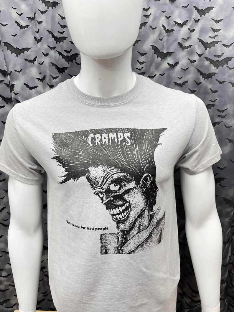 May include: A grey t-shirt with a black and white illustration of a person with a mohawk and the text "CRAMPS" and "Bad music for bad people".