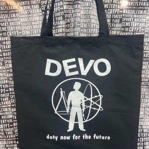 May include: Black tote bag with a white Devo logo and the text "duty now for the future". The logo is a circle with a stick figure holding a protractor and a stylized atom.