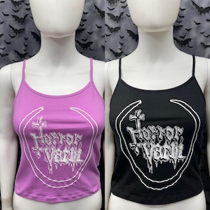 Puede incluir: Dos tops cortos sin mangas con un estampado gráfico de una cruz y el texto "Horror Vacui". Un top es negro y el otro es morado.