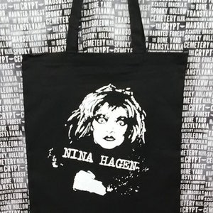 May include: Black tote bag with white text and a black and white image of Nina Hagen. The text on the bag reads "Nina Hagen" and is surrounded by a repeating pattern of words related to Halloween and horror.