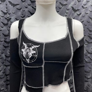 May include: Black ribbed crop top with long sleeves and cut-out shoulders. The top features white stitching and a white graphic of a goat head inside a circle. The top is displayed on a mannequin.