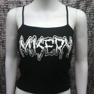 Può includere: Canotta nera con la scritta "MISERY" in bianco, con un font stilizzato e gocciolante. La canotta ha spalline sottili e scollatura rotonda. Lo sfondo è un tessuto fantasia con testo.
