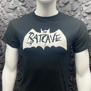 Op de afbeelding: Zwart T-shirt met een witte vleermuis afbeelding en de tekst "The Batcave" in een gestileerd lettertype.