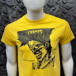 May include: Yellow t-shirt with a black and white illustration of a person with a wild hairstyle. The text "CRAMPS" is above the illustration and the text "...music for bad people" is below the illustration.