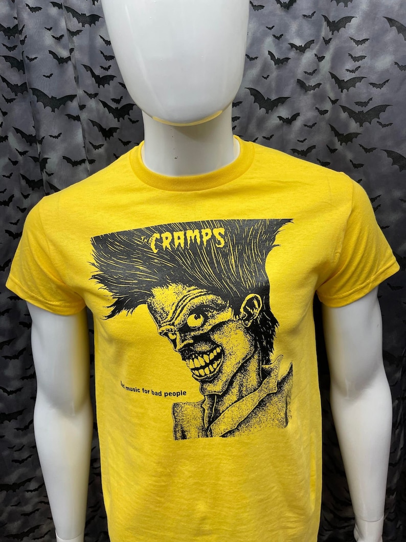 May include: Yellow t-shirt with a black and white illustration of a person with a wild hairstyle. The text "CRAMPS" is above the illustration and the text "...music for bad people" is below the illustration.