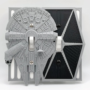 May include: A gray and black light switch cover featuring a 3D printed design of the Millennium Falcon and a Death Star. The switch cover is made of plastic and has a textured surface.