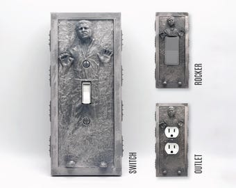 May include: A gray, textured light switch and outlet cover plate set featuring a relief sculpture of a figure encased in a block. The set includes a standard toggle switch plate, a rocker switch plate, and a duplex outlet plate, labeled SWITCH, ROCKER, and OUTLET.