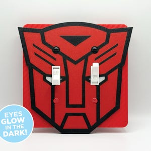 May include: Red and black Transformers-themed light switch cover with two white switches. The design features the Autobot symbol. The text "EYES GLOW IN THE DARK!" is on a blue circle.