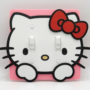May include: A pink light switch cover featuring a white cartoon cat face with a red bow. The cat's face has black outlines, black eyes, and a yellow nose. Two light switches are in the center, with the word "ON" visible.