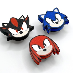 May include: Three colorful, cartoon character face cutouts. The characters are Shadow, Sonic, and Knuckles. Shadow is black, red, and white. Sonic is blue and white. Knuckles is red and white. The cutouts are on a white surface.