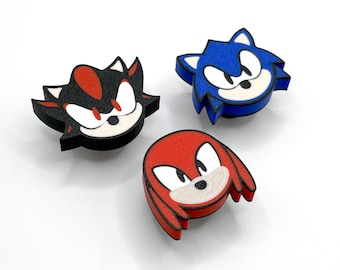 May include: Three cartoon character face cutouts: Shadow, Sonic, and Knuckles. Shadow is black, red, and white. Sonic is blue and white. Knuckles is red and white. The cutouts are on a white surface.