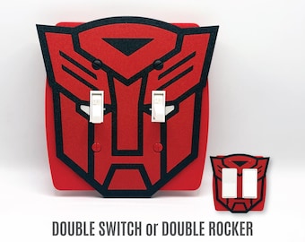 May include: A red and black light switch cover shaped like the Autobot insignia from Transformers. The cover features two white toggle switches or two white rocker switches, with the text DOUBLE SWITCH or DOUBLE ROCKER printed below.