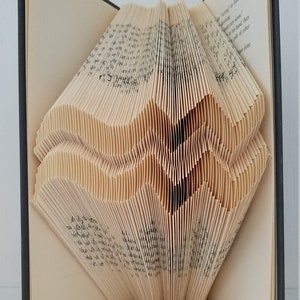 May include: A book art sculpture, folded into the shape of a heart, with the pages fanned out. The book is open and the spine is visible. The book is on a white surface with a small white card with the text "THEBOOKMAGICIAN" on it.