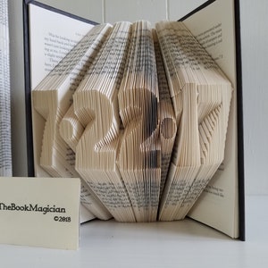 May include: A book art sculpture shaped like the number 2017. The book is open and the pages are folded to create the shape. The book is on a white surface. The Book Magician is printed on a white card in the foreground.