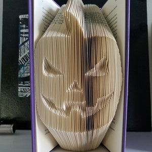 May include: A book folded into the shape of a jack-o'-lantern. The book is open and the pages are folded to create the shape of a face. The book is purple and has the text "TheBookMagician c2018" on the front.