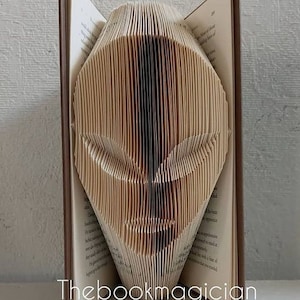 May include: A book art sculpture of an alien face, created by folding the pages of a book. The sculpture is made from a brown book with white pages.