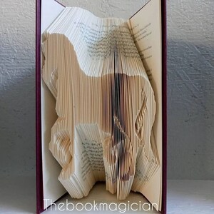 May include: A book art sculpture of a dog made by folding the pages of a book. The book is bound in burgundy and the pages are cream colored. The sculpture is titled "Thebookmagician".
