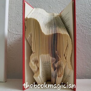 May include: A book art sculpture of a horse, folded from the pages of a book with red covers. The book is standing upright on a white surface. The text "thebookmagician" is printed on the bottom of the book.