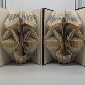 May include: Two books with pages folded to create a three-dimensional sculpture of the scales of justice. The cream-colored paper pages are precisely creased to form the central pillar and balanced pans of the symbol, set against dark book covers.