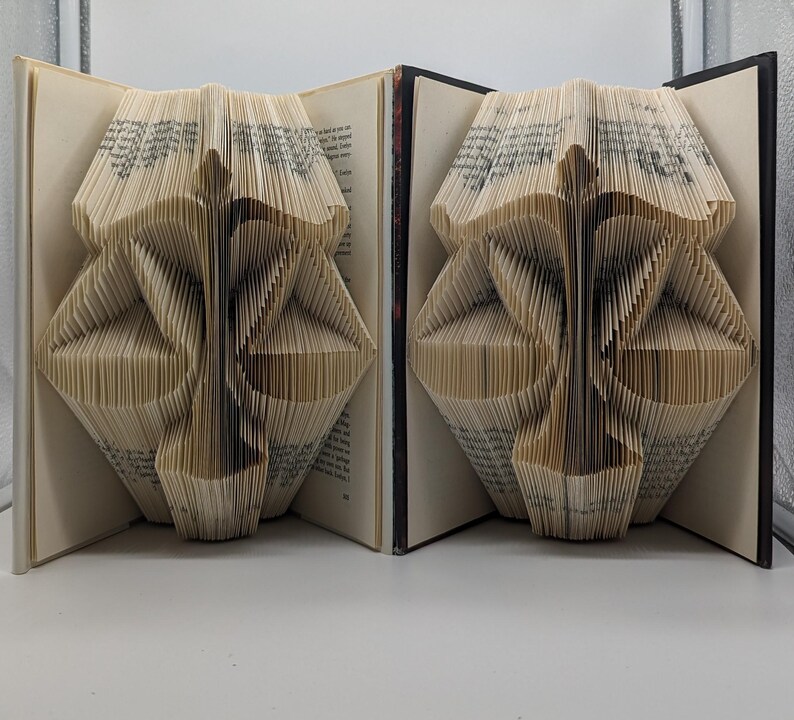 May include: Two books with pages folded to create a three-dimensional sculpture of the scales of justice. The cream-colored paper pages are precisely creased to form the central pillar and balanced pans of the symbol, set against dark book covers.