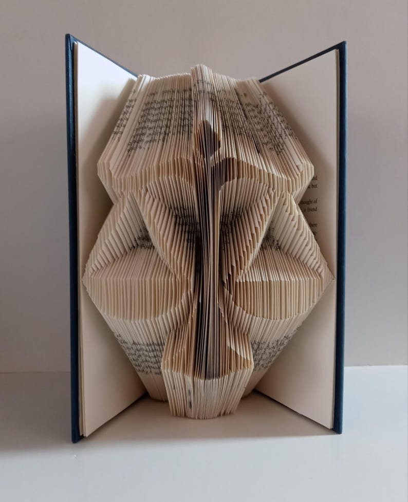 May include: A book with pages folded to create a three-dimensional sculpture of a scale of justice. The book has a dark blue cover and the folded paper pages form the central pillar and two balanced pans of the scale in a vertical orientation.