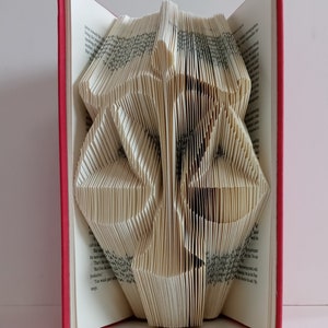 May include: A book with folded pages forming the shape of a scale of justice. The book has a red cover and white pages with black printed text. The pages are precisely creased and manipulated to create a three-dimensional silhouette of a balance scale.