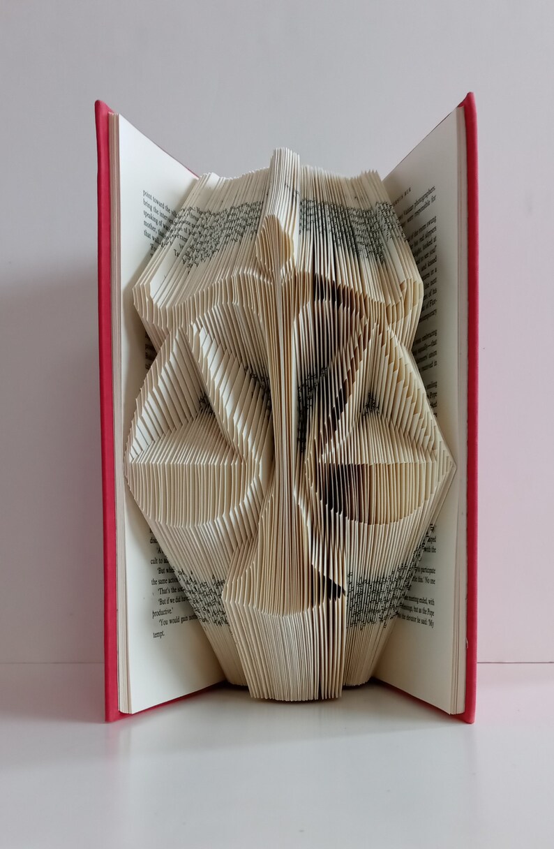 May include: A book with folded pages forming the shape of a scale of justice. The book has a red cover and white pages with black printed text. The pages are precisely creased and manipulated to create a three-dimensional silhouette of a balance scale.