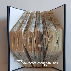 May include: A book art sculpture shaped like the year 2022. The pages of the book are folded to create the numbers. The book is closed and standing upright on a white surface. The book has a black spine and the text "Thebookmagician" is printed on the front cover.