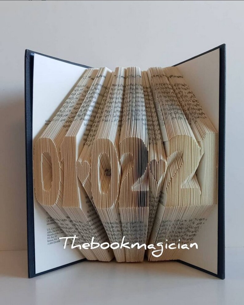 May include: A book art sculpture shaped like the year 2022. The pages of the book are folded to create the numbers. The book is closed and standing upright on a white surface. The book has a black spine and the text "Thebookmagician" is printed on the front cover.