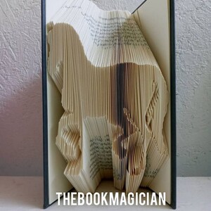 May include: A book art sculpture of a horse made by folding the pages of a book. The book is open and the sculpture is visible. The book has a dark blue cover and the text "THEBOOKMAGICIAN" is printed on the bottom of the book.