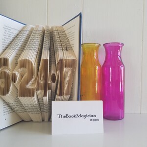 May include: A book with the date 6.24.17 folded into the pages. The book is open and standing upright on a white surface. There are two glass vases, one yellow and one pink, behind the book.