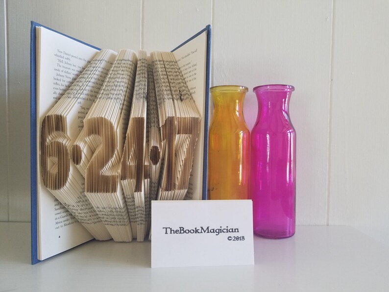 May include: A book with the date 6.24.17 folded into the pages. The book is open and standing upright on a white surface. There are two glass vases, one yellow and one pink, behind the book.