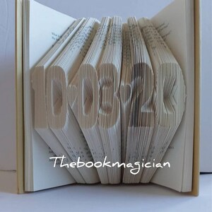 May include: A book art sculpture shaped like the numbers 10.03.2020. The book is folded and the pages are cut to create the shape. The book is open and the spine is visible. The text "Thebookmagician" is printed on the bottom of the book.
