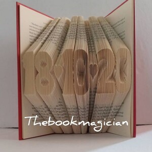May include: A book with the numbers 18 and 22 folded into the pages, with a heart symbol between them. The book is red and the pages are white. The text "Thebookmagician" is printed on the bottom of the book.