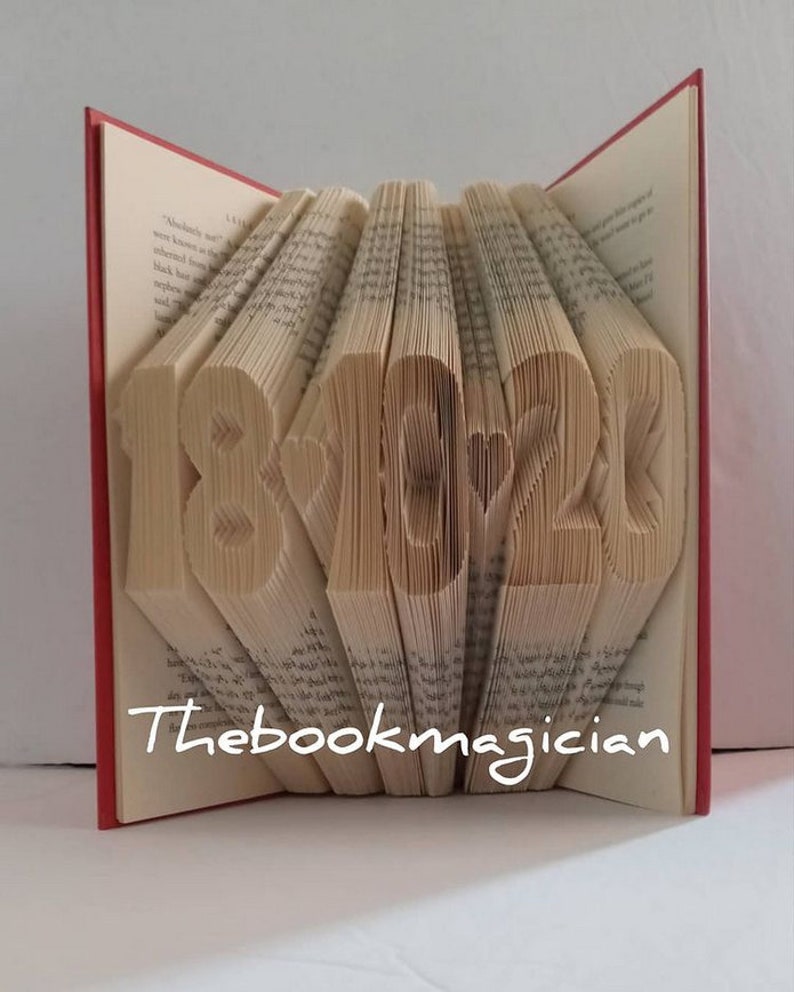 May include: A book with the numbers 18 and 22 folded into the pages, with a heart symbol between them. The book is red and the pages are white. The text "Thebookmagician" is printed on the bottom of the book.
