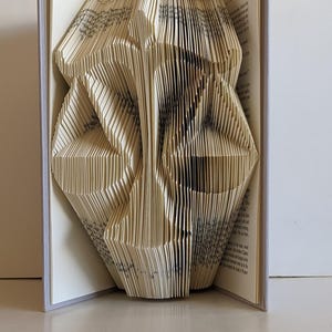 May include: A book art sculpture created from a hardcover book with pages folded to form the shape of a scale of justice. The off-white paper pages are precisely creased to create a three-dimensional silhouette of a balanced legal scale.