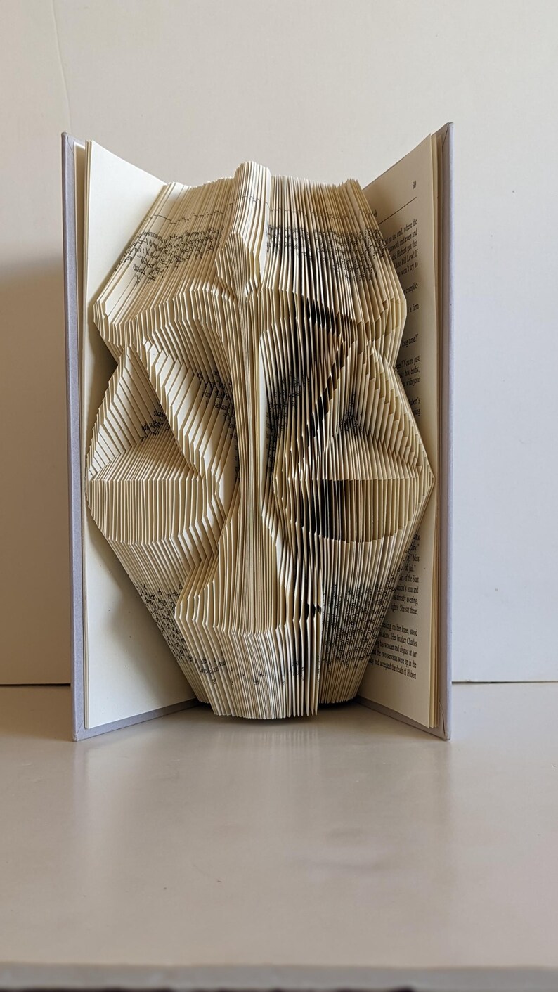 May include: A book art sculpture created from a hardcover book with pages folded to form the shape of a scale of justice. The off-white paper pages are precisely creased to create a three-dimensional silhouette of a balanced legal scale.