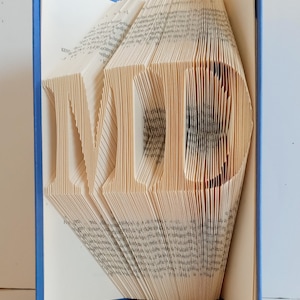 May include: A book with pages folded to create the letters "MD" in a 3D design. The book has a blue cover.