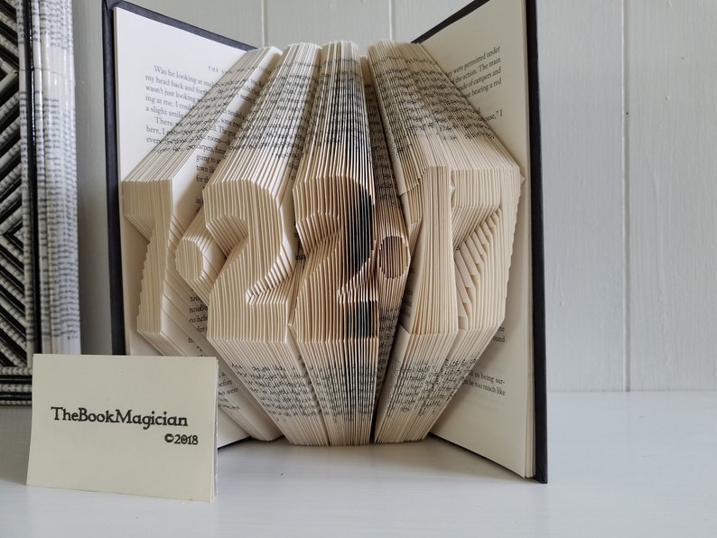 May include: A book art sculpture shaped like the number 2017. The book is open and the pages are folded to create the shape. The book is on a white surface. The Book Magician is printed on a white card in the foreground.