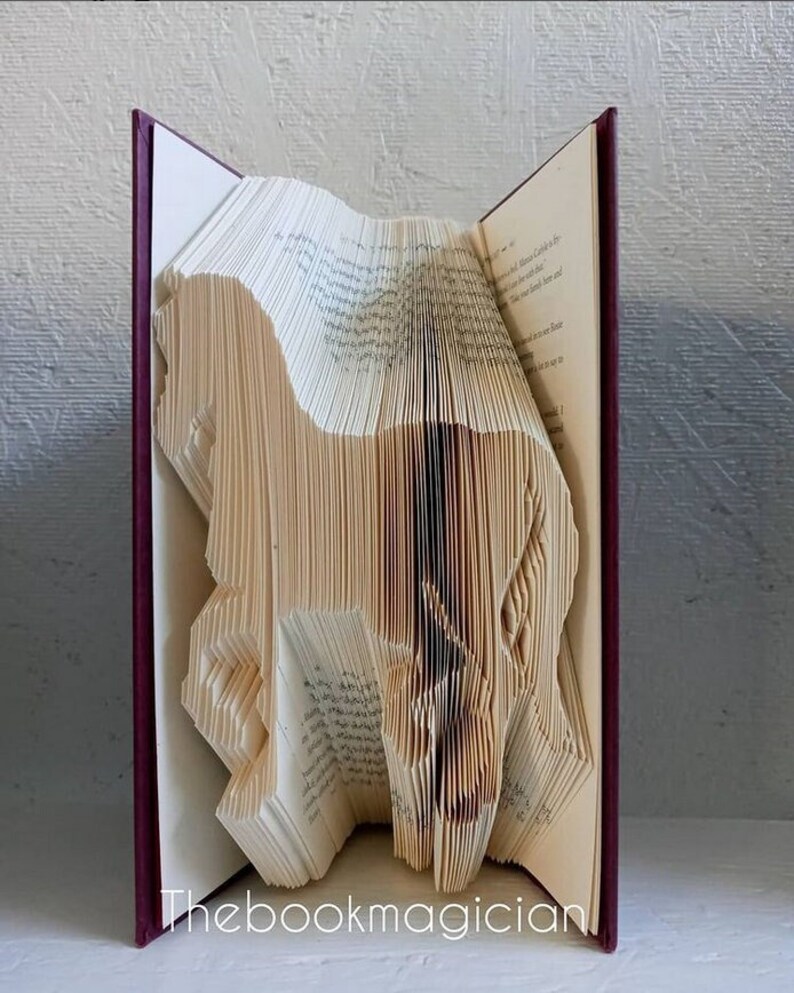 May include: A book art sculpture of a dog made by folding the pages of a book. The book is bound in burgundy and the pages are cream colored. The sculpture is titled "Thebookmagician".