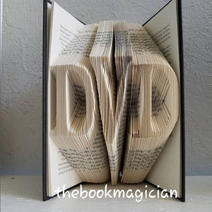 May include: A book art sculpture of the letters "DVD" created by folding the pages of a book. The book is open and the pages are folded to create the letters. The book is closed on the right side and the spine is black.